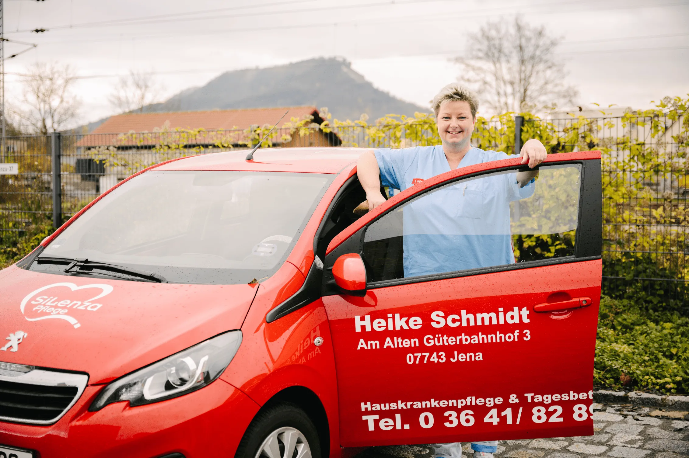 Pflegekraft von Silenza lehnt lächelnd an der offenen Tür eines roten Dienstwagens, im Hintergrund ein Berg bei Jena
